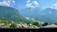 Panoramic view from a balcony with mountains, green valley, village houses, and clear blue sky