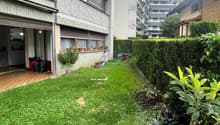 Building with tiled entrance, open garage, small garden area, balcony with potted plants, hedge, grass lawn, trash bins, and a tool cart.
