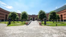 A modern architectural building made of red bricks with multiple windows, wide courtyard with trees and paved entrance