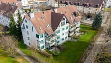 Aerial view of a multi-story residential building with brown roofs, white walls, and green window frames. Balconies are visible on the upper floors. The building is surrounded by a well-maintained green lawn and several trees. A car is parked in the parki