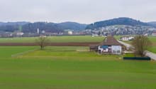 The image shows a rural countryside landscape with rolling green hills, a small village in the distance, and a farmhouse with a balcony or terrace in the foreground. The scene has a peaceful, pastoral feel with fields, fences, and a few trees dotting the