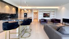 modern kitchen with marble countertop, wooden cabinets, 4 bar stools, living room with couch, 2 armchairs, TV, wooden floor