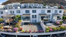 White multi-story building with multiple balconies, plants, flowers, cars parked in the ground floor parking, tables and chairs outside