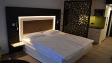 Modern bedroom with a large bed, white bedding, and a decorative black and white patterned wall panel. The room also features recessed lighting in the headboard and a fireplace or heating unit on the wall.