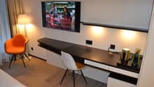 Modern living room with a large TV mounted on the wall, a dark wooden TV stand with drawers, an orange accent chair, and a white chair at a desk area. The room has hardwood flooring and curtains covering a window or balcony door.