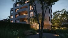 A modern, multi-story apartment building with balconies, surrounded by trees and a well-landscaped garden area with a bench. The building has a sleek, contemporary design with large windows and a mix of materials like wood and concrete.