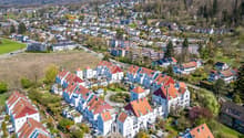Aerial view of a residential area with many houses, some with red roofs, surrounded by trees and greenery.