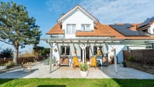 white house, red roof, solar panels, patio with wooden chairs, potted plants, wooden fence, lawn
