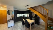 Dining area with a wooden table and black chairs, wooden staircase, kitchen with black cabinets, round mirror, and a wooden door.