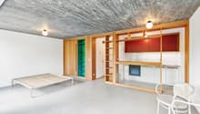 Open-concept studio apartment with concrete ceiling, wooden built-in shelving and cabinets, a bed frame, and a white chair. The room has a minimalist and modern design aesthetic.