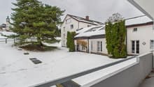 Two-story building, white exterior, covered in snow, large front yard, snow on ground, trees, small solar panels, fenced yard, another building in the background.