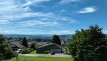 The image shows a scenic view of a residential area surrounded by lush greenery and mountains in the background. The sky is clear with wispy clouds, creating a picturesque setting. The houses in the foreground have a mix of architectural styles and are ne