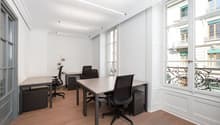 Spacious office space with large windows, hardwood floors, and modern black desks and chairs. The room has ample natural light and a view of the surrounding buildings outside.