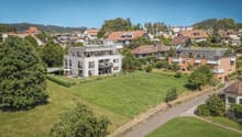Modern apartment building with balconies, located in a residential area with greenery and mountains in the background.