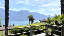 The image shows a scenic outdoor landscape with a view of a lake surrounded by mountains. There is a wooden railing or fence in the foreground, with lush greenery and palm trees in the middle ground. The background features a clear blue sky with some clou