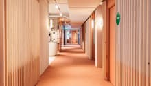 The hallway has wooden slat walls, a peach colored floor, and bright white lighting on the ceiling. At the end of the hallway is a kitchen area with wooden shelves, white tiles, and an island.