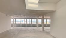 Large open-plan office space with concrete floor, white walls, and large windows providing a view of the surrounding area. The space has a high ceiling with a skylight, allowing natural light to flood the room.
