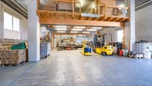 Empty warehouse with a large open area, wooden mezzanine with storage boxes, a few stacks of wooden pallets with boxes, two forklifts, machinery, metal barrels, and containers