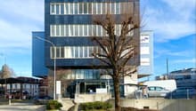 Modern office building with blue exterior, multiple windows, entrance with glass doors, street light, cars parked in front, bicycles parked, small tree in front, signboard with text 'safe' and 'ARMEDIZIN'.