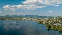 Aerial view of a scenic lakeside town surrounded by mountains and hills, with sailboats on the calm blue waters of the lake and a partly cloudy sky overhead.