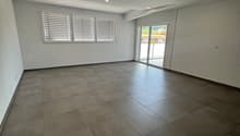 Spacious living room with large windows, gray tile flooring, and recessed lighting. The room has access to a balcony or terrace through sliding glass doors.