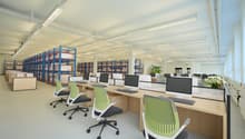 Modern office space with rows of ergonomic chairs, wooden desks, multiple monitors, keyboard and mouse, blue storage racks with boxes, artificial plants and ceiling lights.