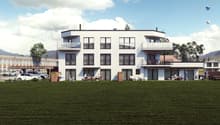 3-story modern apartment building with white exterior, large windows, and balconies. There is a grassy lawn in the foreground and parked cars visible in the background.