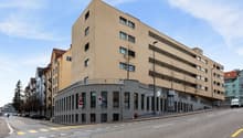 Modern multi-story building with beige exterior, glass windows, and a curved section at the base.