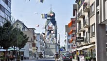 City street with buildings, church, flags, trees, cars, bicycles, street lights, signs, and potted plants.