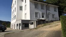 2-story white building with a slanted roof, windows, and a balcony. There is a gravel parking area in front of the building.
