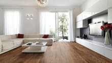 Spacious living room with hardwood flooring, large windows providing natural light, a white sectional sofa, a modern white coffee table, and a decorative chandelier overhead.