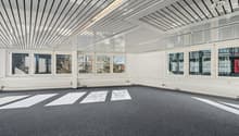 A large empty room with carpeted floor, multiple large windows, metal ceiling panels, recessed lighting, and no furnishings