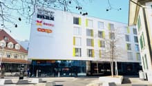 Modern white building with yellow accents, large glass windows, multiple floors, entrance with glass doors, signage for 'REX MAX' and 'COOP', concrete planters, street lights, and a tree in front.