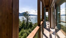 Wooden balcony with a panoramic view of a lake surrounded by mountains and trees. The balcony has large windows and a railing overlooking the scenic landscape.