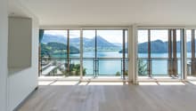 Spacious living room with large windows offering a panoramic view of a lake surrounded by mountains. The room has a minimalist design with light-colored hardwood floors and white walls. The windows provide abundant natural light and a serene, picturesque