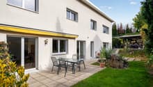 Beige house, large windows, sliding doors, paved patio with table and chairs, awning, garden with plants
