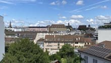 The image shows a panoramic view of a city skyline. The scene is dominated by a blue sky with fluffy white clouds. The cityscape is composed of a mix of residential and commercial buildings, with some greenery visible in the foreground. The buildings have