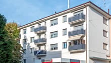 The image shows a multi-story apartment building with several balconies visible on the facade. The building has a modern, clean design with white walls and gray balcony railings. There is a commercial space on the ground floor, with the sign 'Auto Gambino