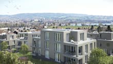 Multi-story apartment building with modern, gray exterior and large windows. The building has balconies on some units. The surrounding area includes a hilly, forested landscape with a small town or village in the distance.