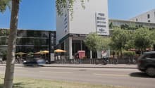 Building with vertical gardens, white walls, multiple signs indicating businesses. Umbrellas, bikes, and cars visible on the street. Trees line the sidewalk, and there are parked cars and bikes on the street.