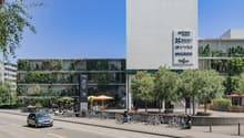 Multi-story building, vertical garden, parking area, street view, outdoor seating area, bikes parked on the side, car driving, trees and greenery, multiple shops on the ground floor, white walls and signs.