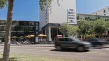modern multi-story building with retail shops on the ground floor, outdoor seating with umbrellas, bicycles parked, sign indicating presence of restaurants and cafes, traffic in the foreground