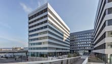 Modern office building with glass facade, multiple floors, and a landscaped area in front.