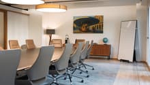 spacious room, white walls, large wooden table, multiple modern chairs, lamp, wooden desk, painting, whiteboard, rug