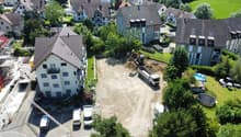Aerial view of a residential area with a construction site in the center. The site has an excavator and a dump truck, indicating ongoing construction work. The surrounding buildings appear to be multi-story residential apartments with tiled roofs. There a