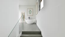 Bathroom with white bathtub, minimalist interior design, grey tiled flooring, glass railing, small window.