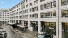 White building with multiple windows, ground floor with shops, parking area in front