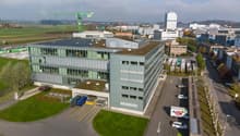 Office building, 4 stories, large windows, grey and white exterior, solar panels on the roof, parking area with cars