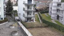 Aerial view of an apartment building in an urban setting with trees, a slope, and other buildings in the distance
