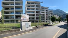 two multistory apartment buildings, white exterior, glass balconies, sidewalk, chain link fence, grassy field, mountain